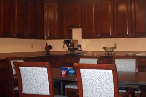 Dining area with a wooden table and upholstered chairs in front of dark wood cabinets and a countertop holding a coffee maker.