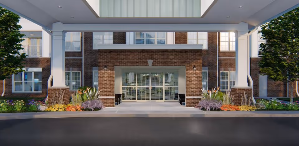 Brick building front entrance with glass doors, white columns, and colorful landscaping.