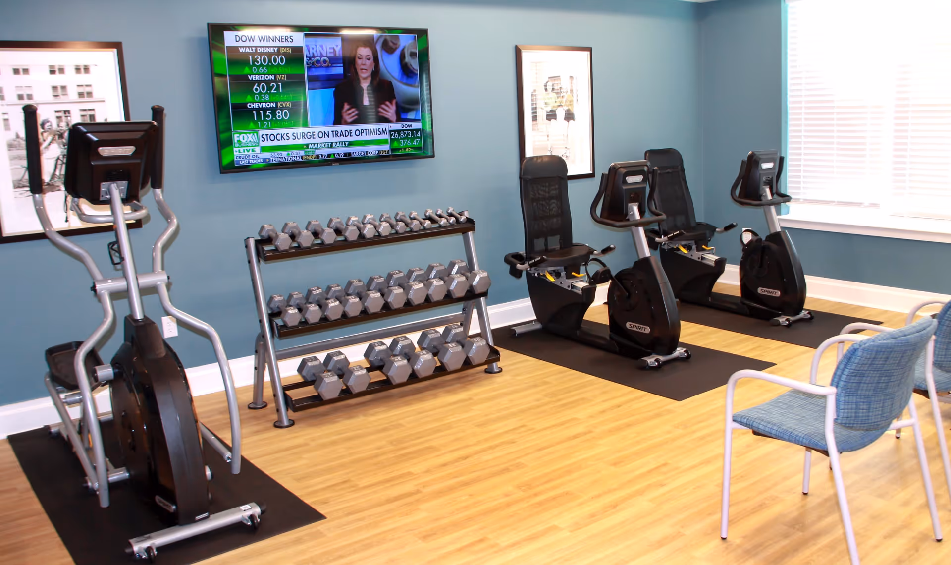 Indoor fitness room with exercise equipment including an elliptical machine, two recumbent bikes, and a rack of dumbbells. A flat-screen TV is mounted on the blue wall showing a news channel. There are two framed pictures on the walls and a window with white blinds letting in natural light. A blue cushioned chair with white metal frame is visible in the foreground.