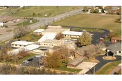 Aerial view of a healthcare facility complex with multiple connected buildings, parking areas, surrounding trees, and open grassy fields.