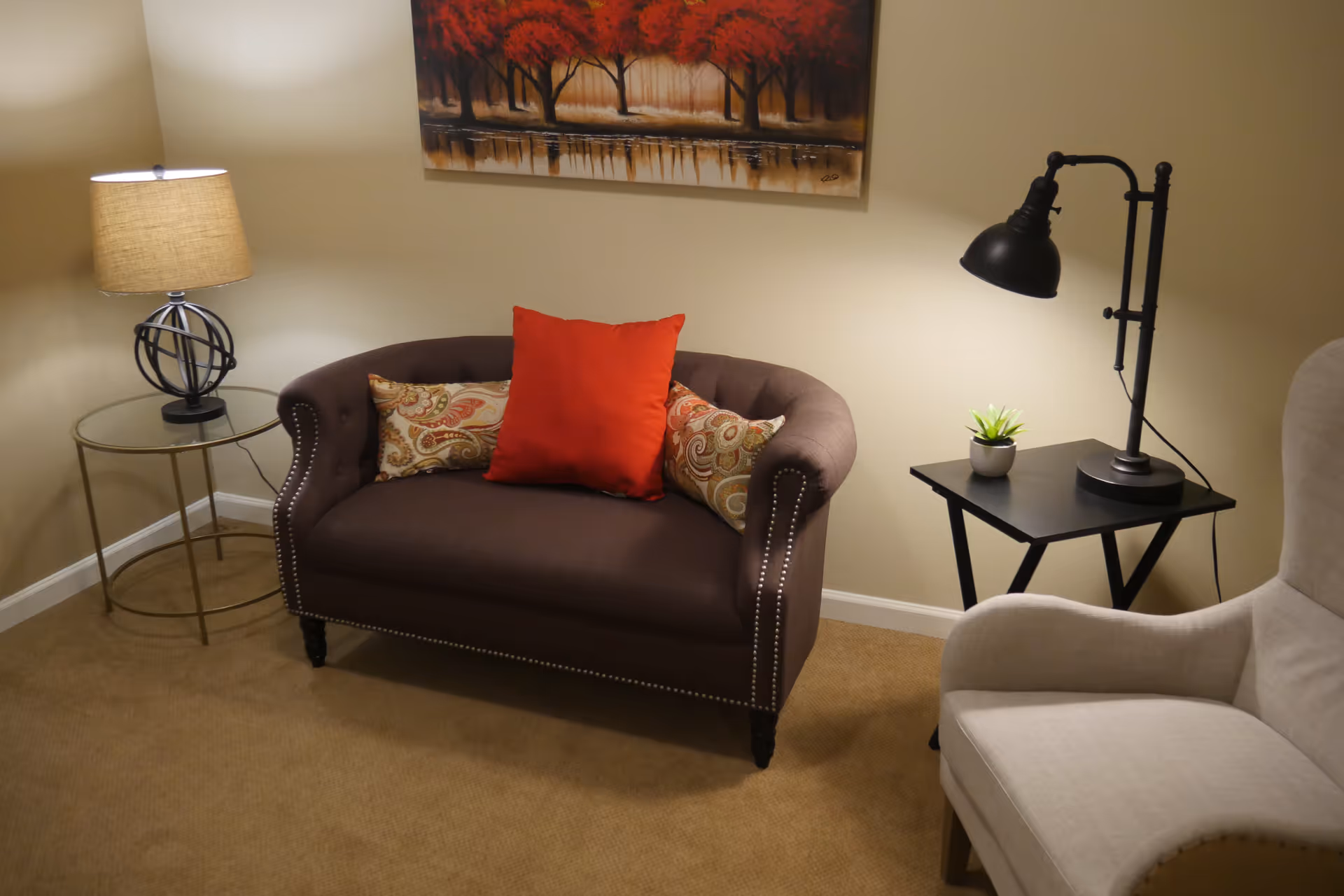 Cozy sitting area with a brown loveseat topped by a red accent pillow, side tables with lamps, and a beige armchair.