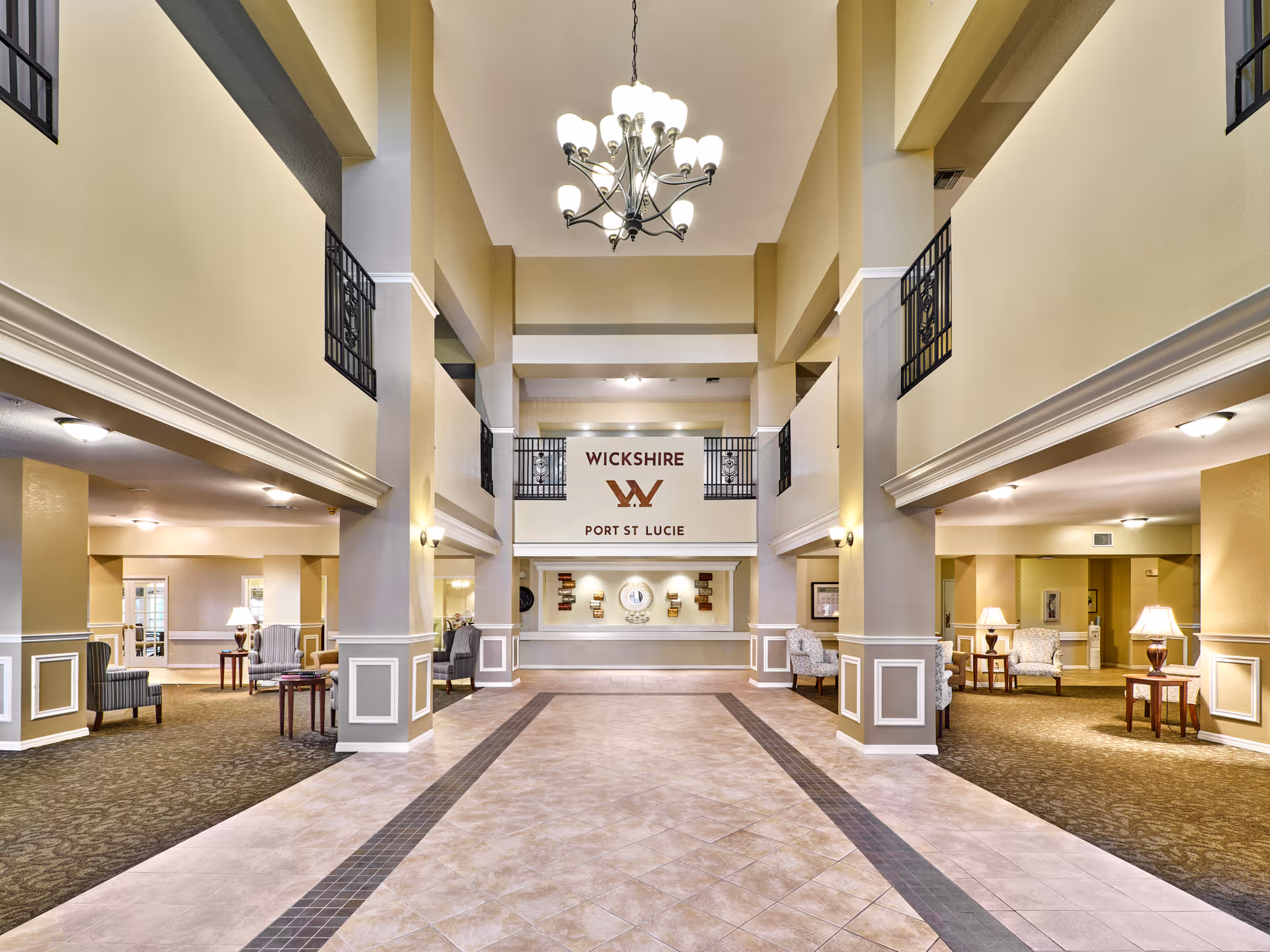 Spacious and well-lit lobby area of an assisted living facility with high ceilings, a chandelier, and beige walls. The floor is tiled in the center with carpeted seating areas on both sides furnished with armchairs and small tables with lamps. A sign on the far wall reads 'WICKSHIRE PORT ST LUCIE'.