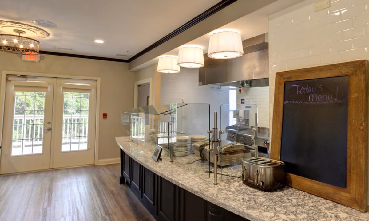 Interior view of a senior living facility dining area with a serving counter featuring a glass sneeze guard, plates stacked, a toaster, and a chalkboard sign that reads 'Today's Menu!'. There are three hanging lamps above the counter and double glass doors leading outside.