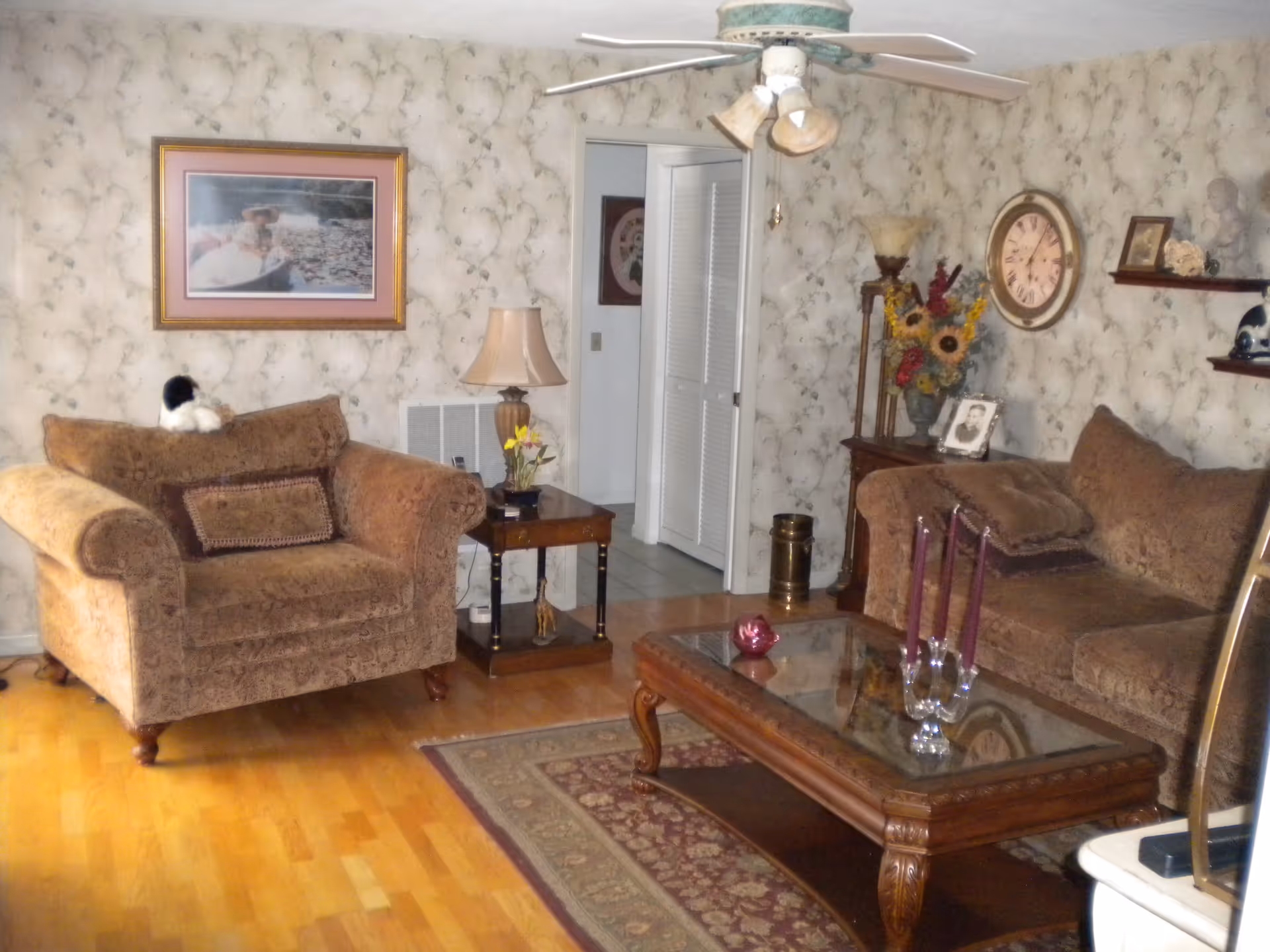 A cozy living room with two brown upholstered sofas, a wooden coffee table with a glass top, and a patterned area rug. The walls have floral wallpaper and are decorated with a framed picture, a large clock, and shelves holding various decorative items. A ceiling fan with lights hangs above, and there is a side table with a lamp and flowers next to one sofa.