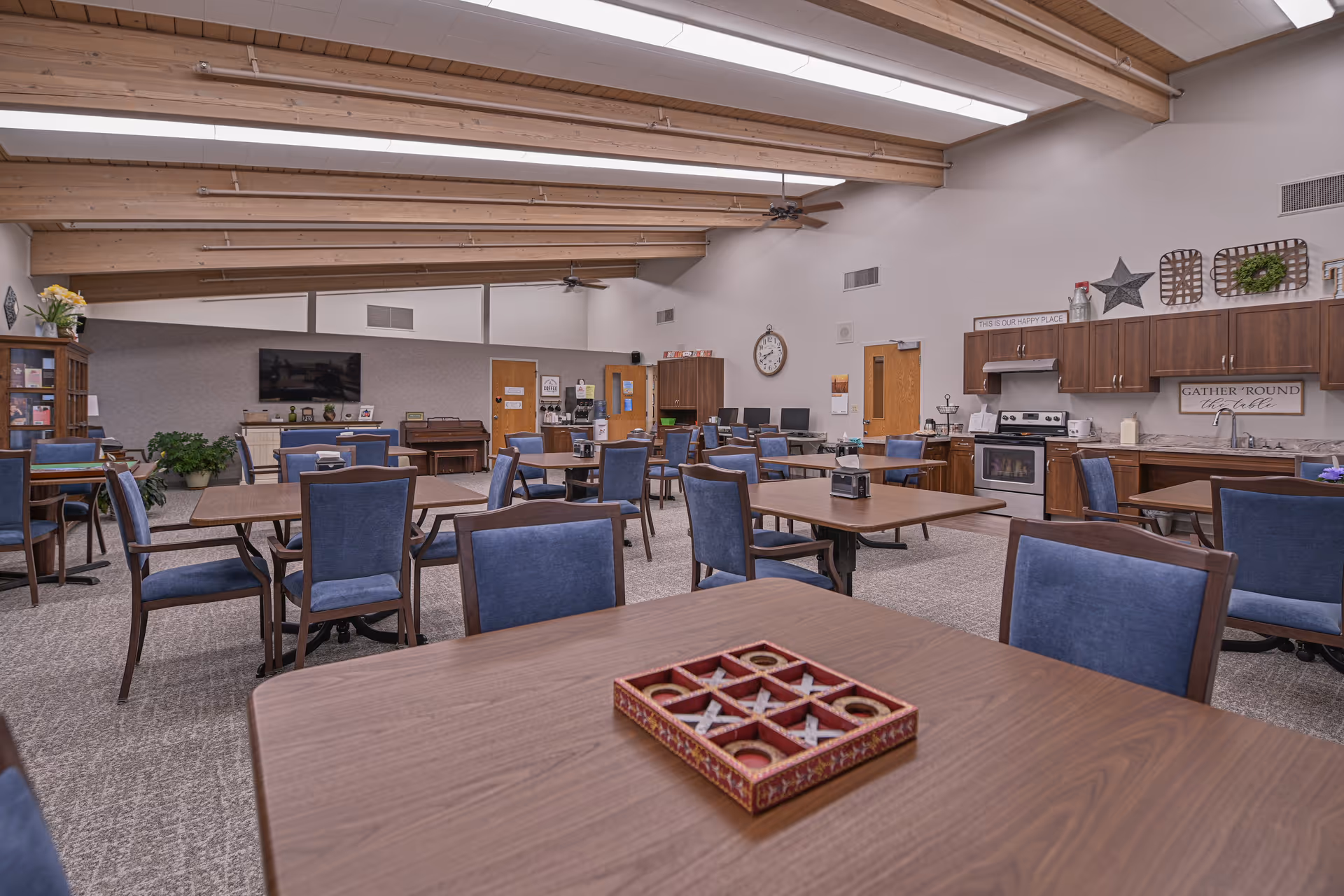 A spacious common area in a senior living facility with multiple wooden tables and blue cushioned chairs arranged throughout the room. The room features a high ceiling with exposed wooden beams and ceiling fans. On the right side, there is a kitchen area with wooden cabinets, a stove, and a sink. The walls are decorated with signs and wreaths. In the background, there is a TV mounted on the wall, a piano, and some computer stations. The room is well-lit with natural and artificial light.