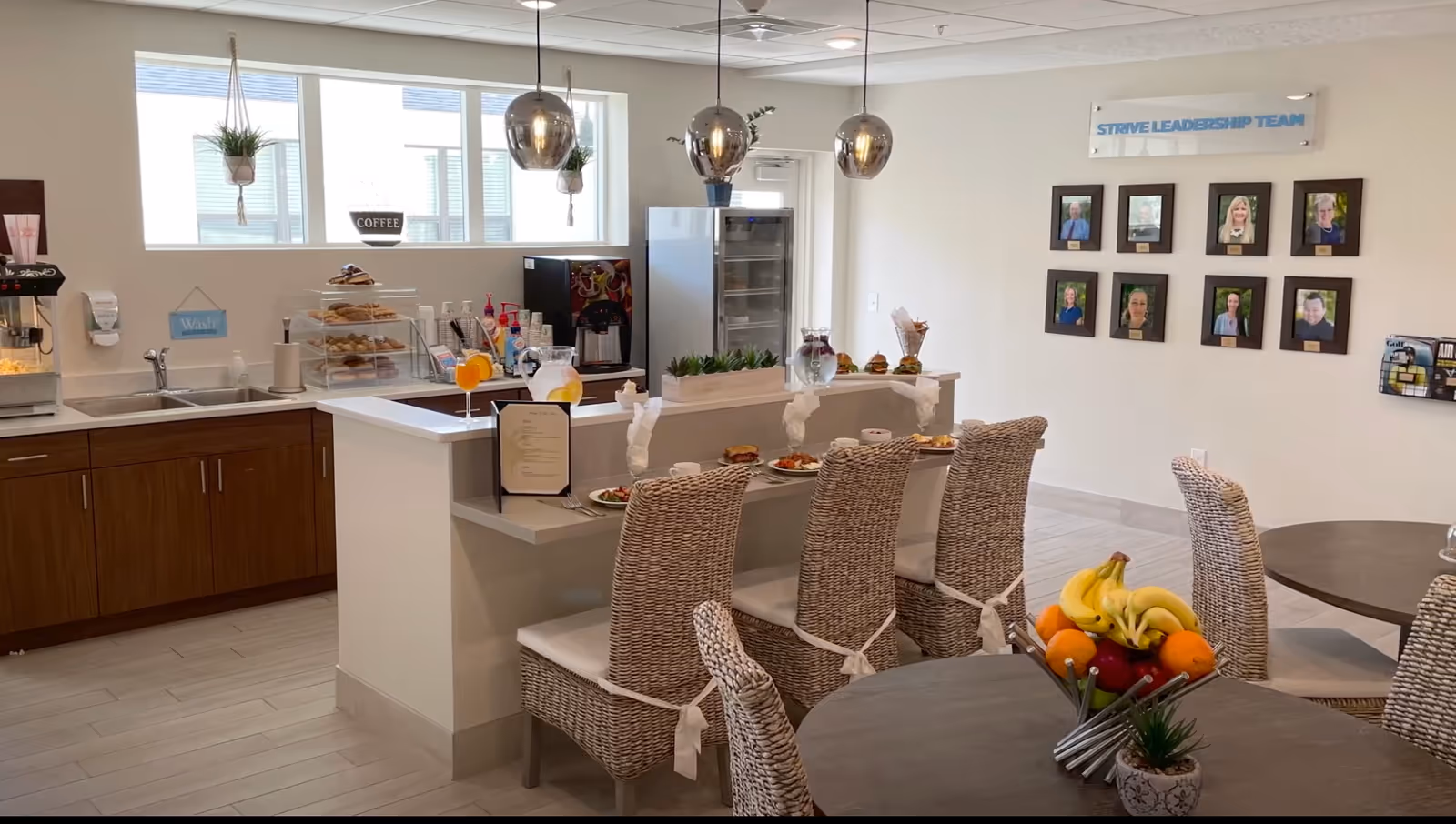A bright and modern dining area in a senior living community featuring a counter with four wicker chairs set with plates of food and drinks. Behind the counter is a kitchen area with a sink, coffee machine, and a display case with pastries. On the wall, there are framed photos labeled 'Strive Leadership Team'. A round table with wicker chairs and a fruit bowl is in the foreground.