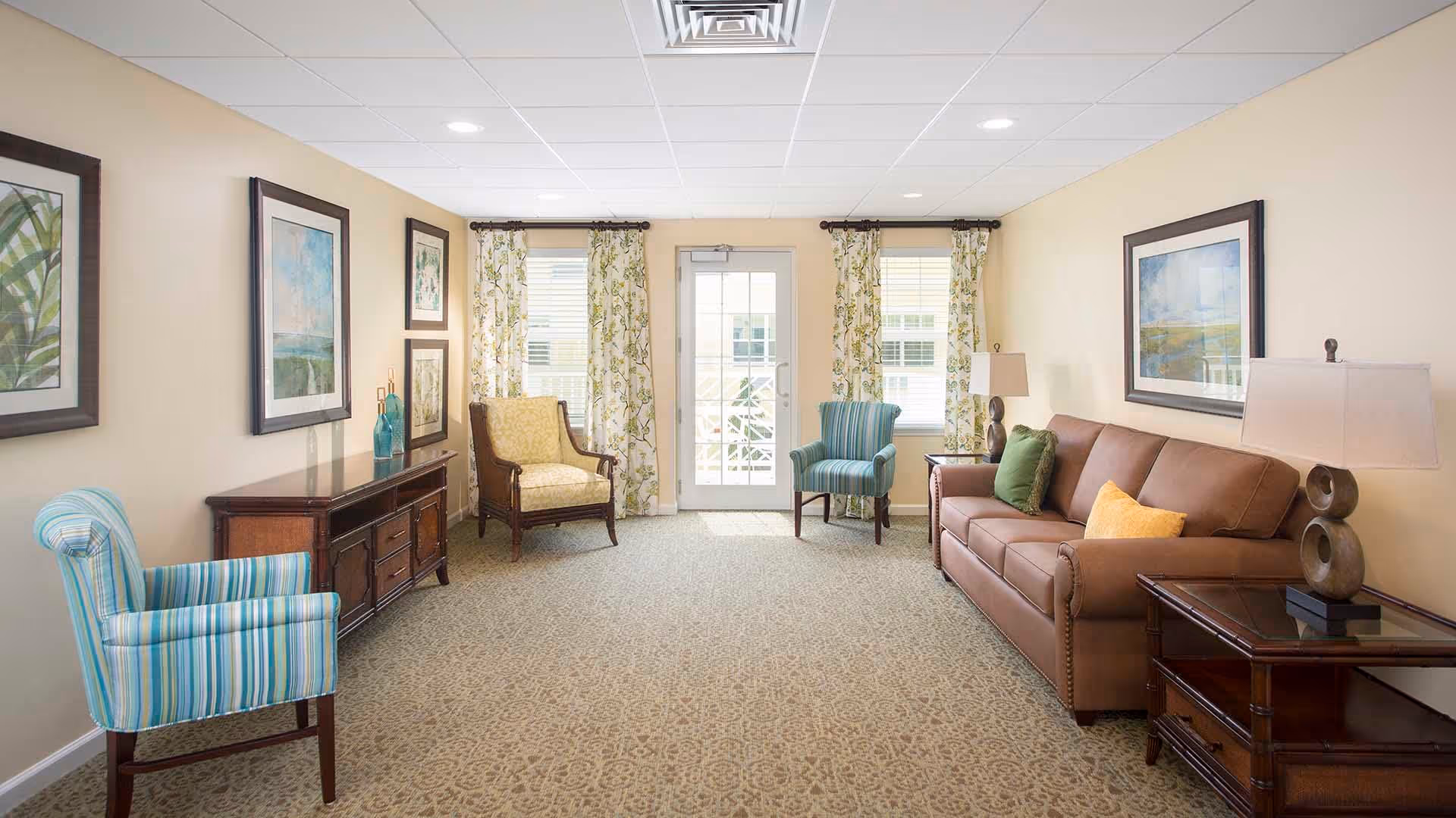 A bright and cozy living room with a brown leather sofa adorned with green and yellow pillows, two striped blue armchairs, and a yellow patterned armchair. The room features beige walls decorated with framed artwork, floral curtains on windows flanking a glass door, and wooden side tables with lamps.