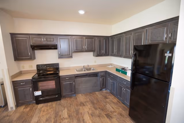 L-shaped kitchen with dark wood cabinets, black appliances (stove and refrigerator), a double sink, and wood-look flooring.