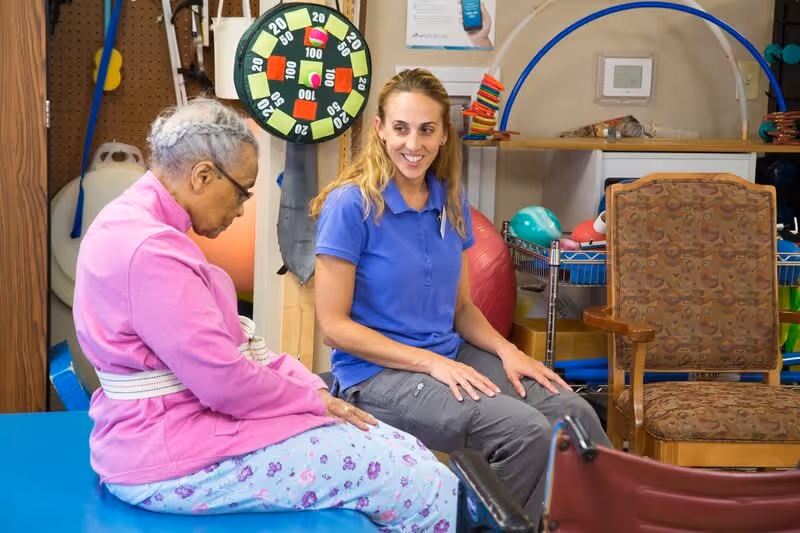 An elderly woman in a pink jacket and floral pants sits on a blue therapy table with a white belt around her waist, while a smiling female caregiver in a blue polo shirt and gray pants sits beside her in a room with various therapy and exercise equipment.