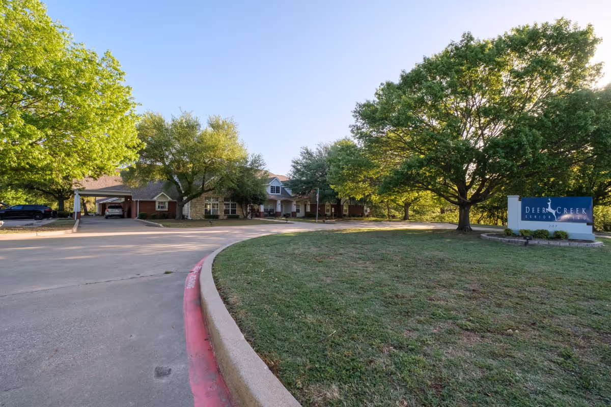Front driveway, lawn and entrance of Deer Creek Senior Living with trees and a facility sign.