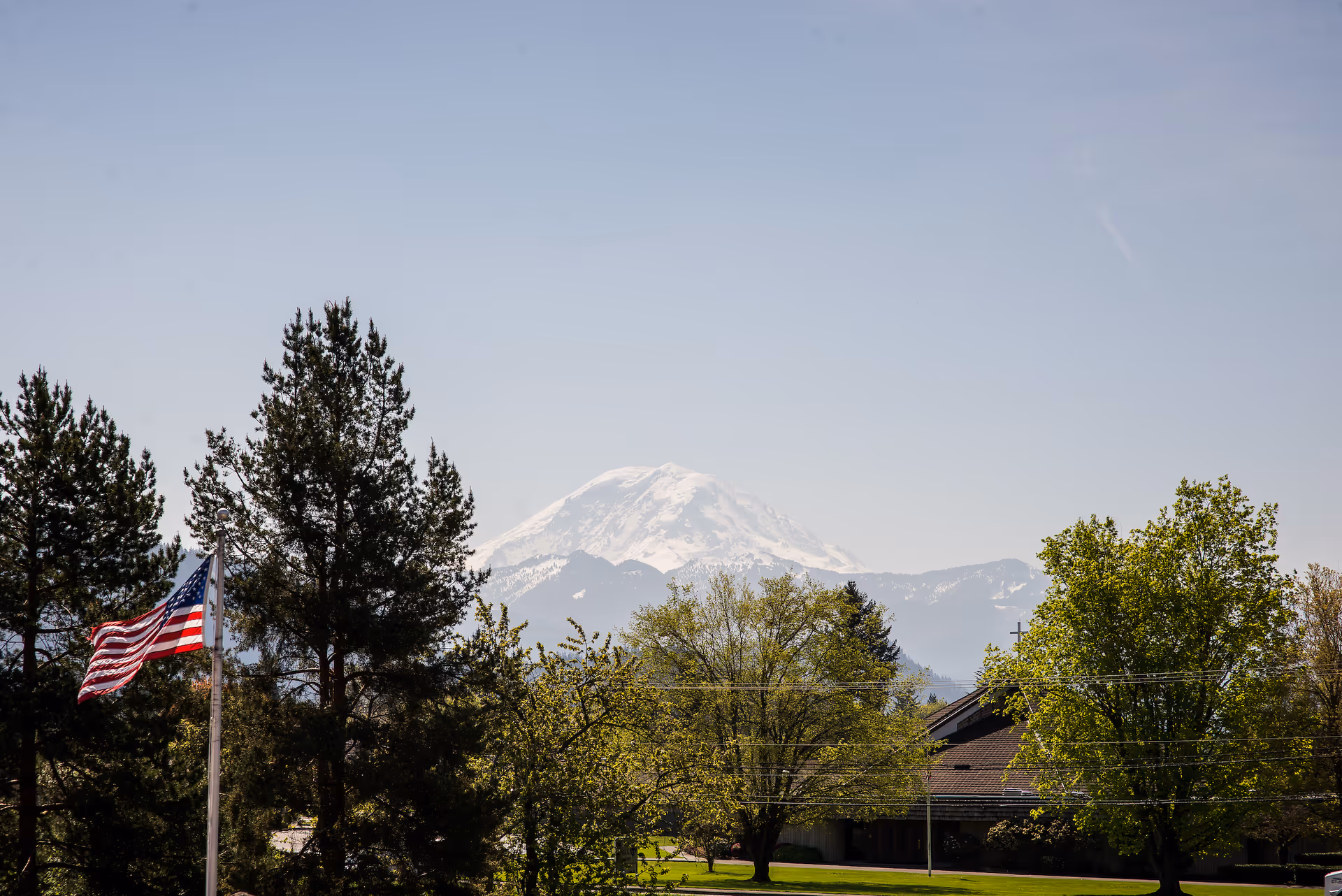 Snow-capped mountain seen over trees with an American flag on a pole and a building roof under a clear sky.