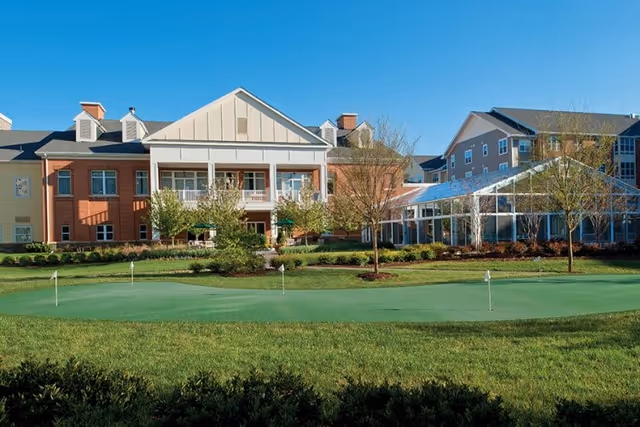 Exterior view of Ashby Ponds senior living facility with a well-maintained putting green in the foreground, surrounded by green grass and small trees, under a clear blue sky.