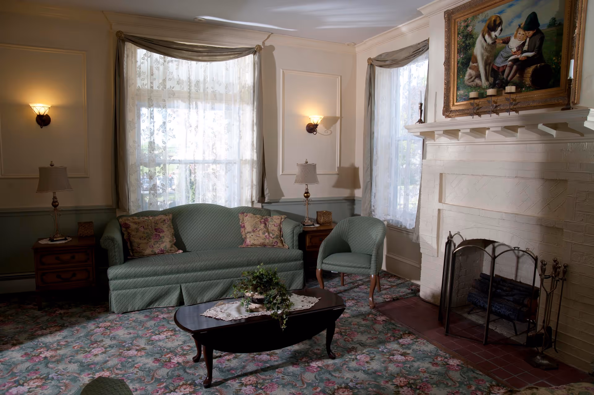 A cozy living room with a green upholstered sofa and matching armchair, floral patterned carpet, wooden coffee table with a lace doily and plant, two side tables with lamps, a white brick fireplace with a metal screen and fireplace tools, and a large framed painting above the mantel depicting children and a dog. Sheer floral curtains cover the windows.