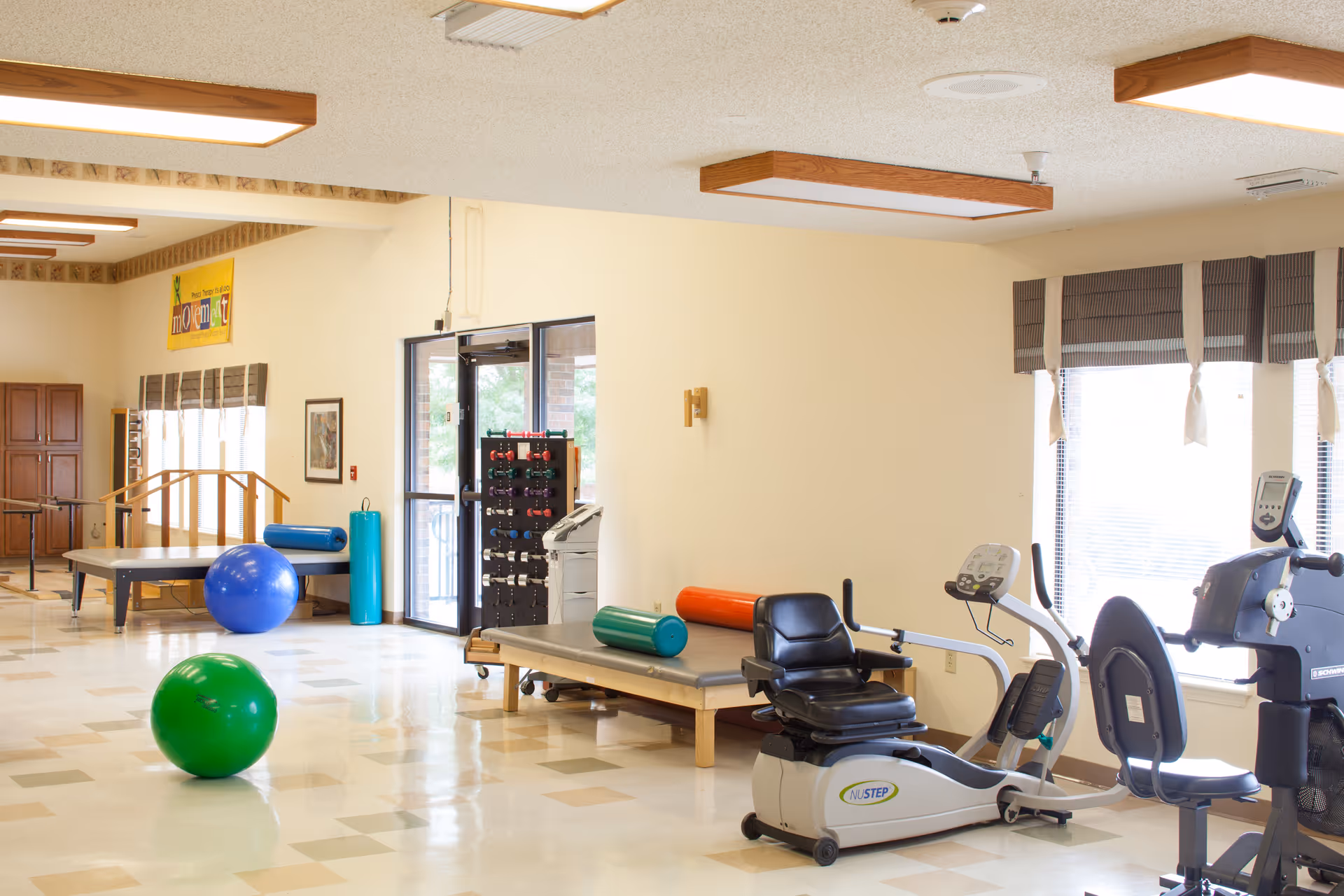 Bright rehabilitation gym with exercise bikes, therapy tables, large exercise balls, and weights.