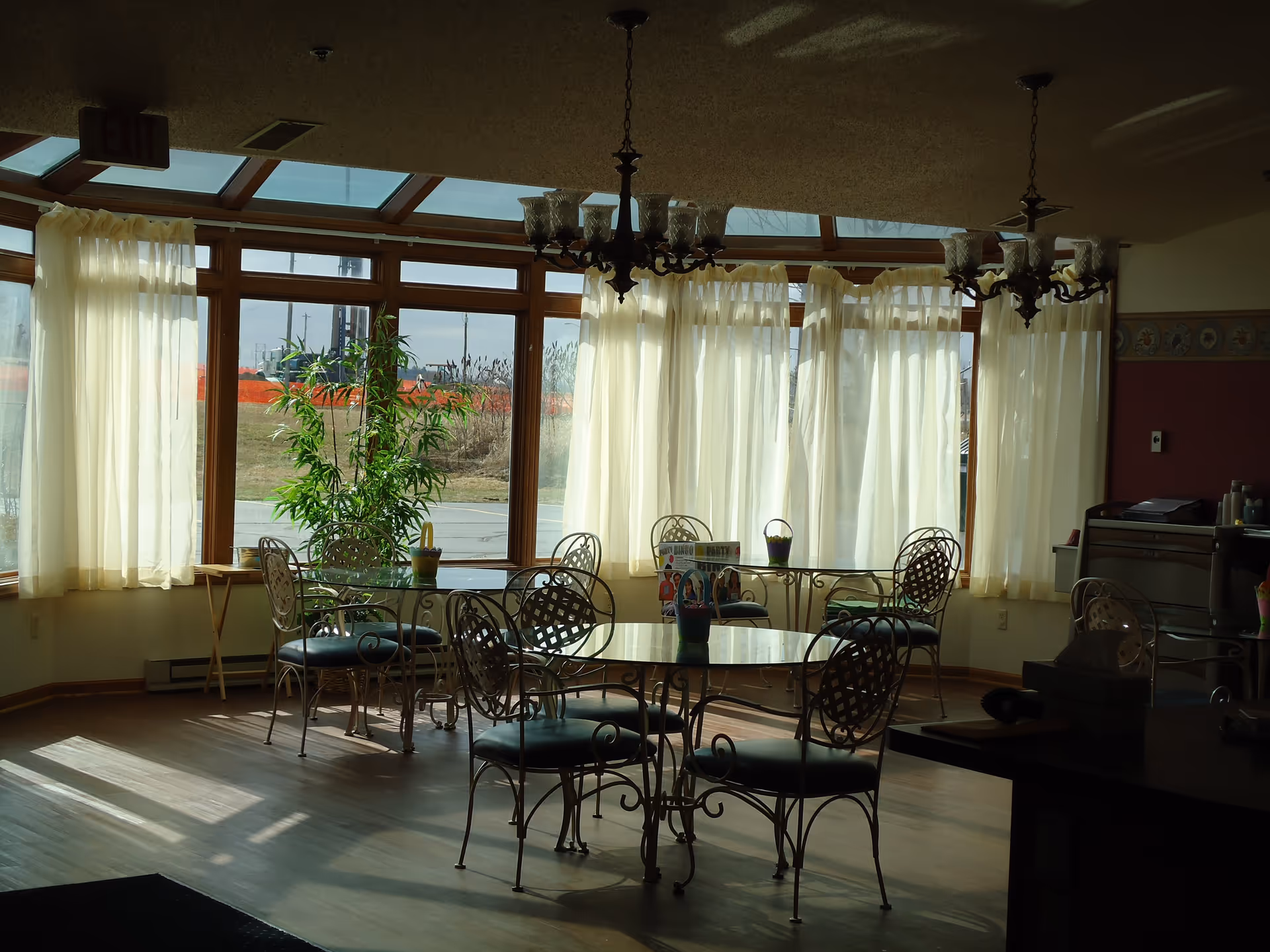 Sunlit communal dining area with round glass tables and wrought-iron chairs in front of large curtained windows.
