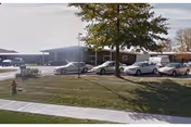 Single-story brick facility building with a front lawn, a tree, parked cars and a sidewalk.