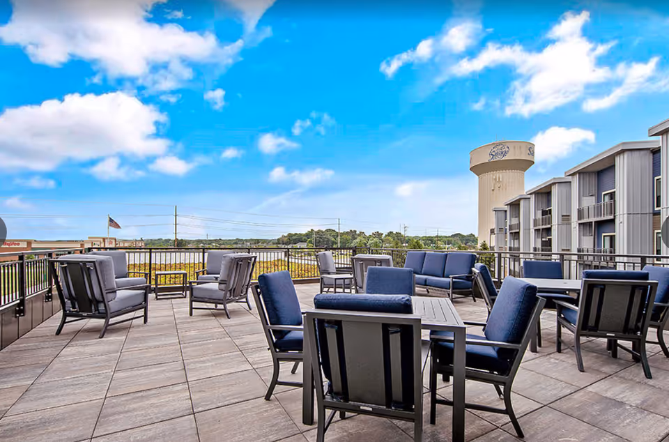 Outdoor patio area with multiple seating arrangements including cushioned chairs and sofas under a bright blue sky with scattered clouds. A water tower and a multi-story building are visible in the background.