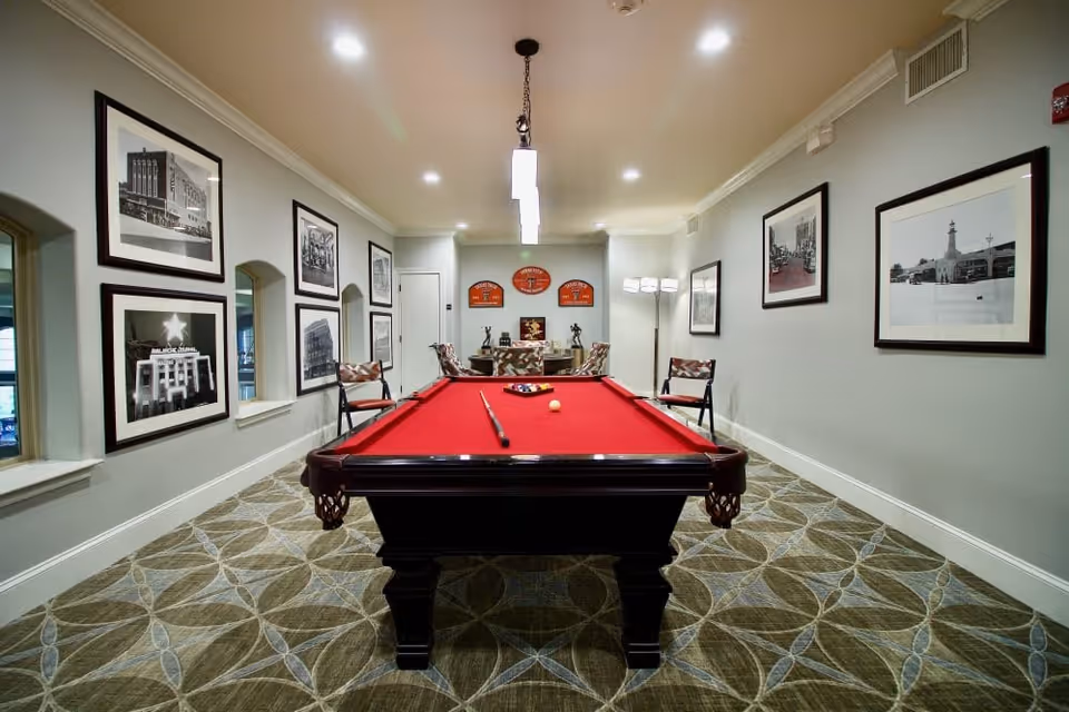 Interior room with a red felt pool table in the center, surrounded by chairs. The walls are decorated with framed black and white photographs and memorabilia. The room has patterned carpet flooring and modern ceiling lights.