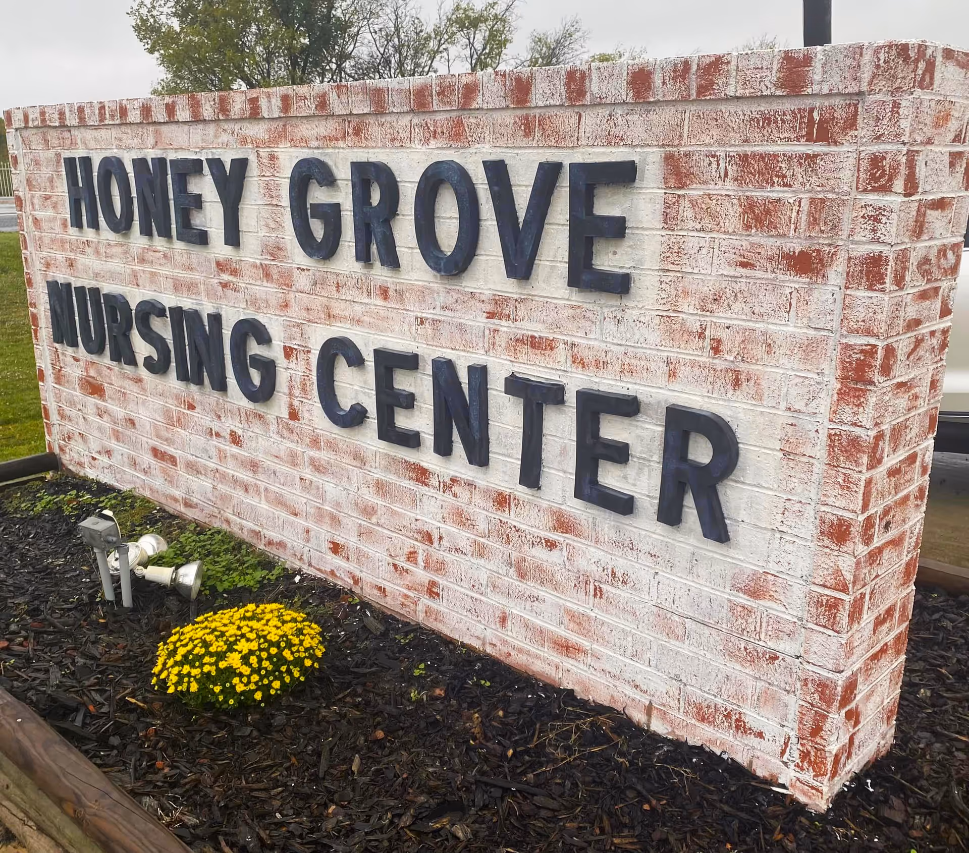 A brick sign with black raised letters that reads 'HONEY GROVE NURSING CENTER' surrounded by mulch and a small yellow flowering plant in front.