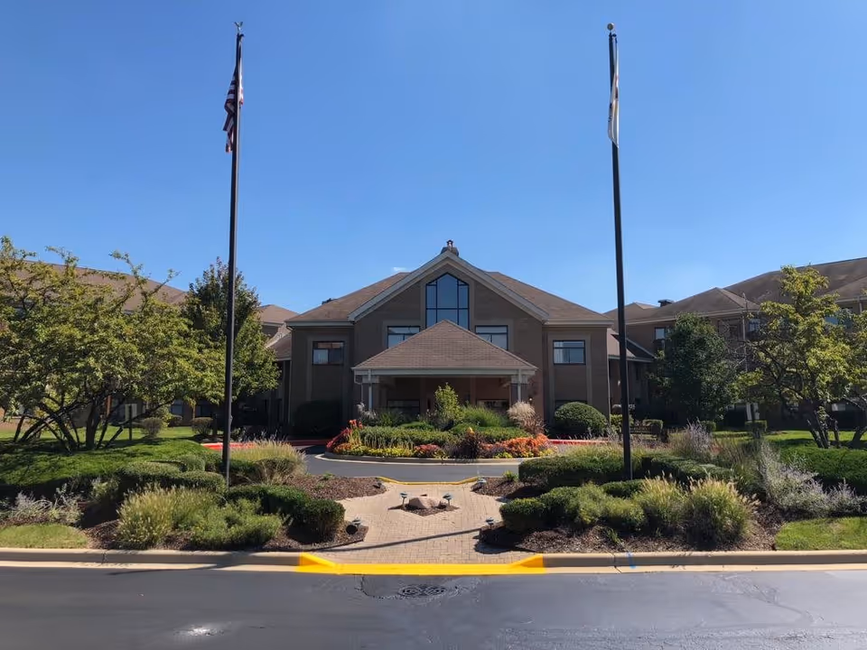 Front exterior view of Bridgeway of Bensenville Supportive Living building with a peaked roof entrance, two flagpoles with flags, landscaped garden with bushes and flowers, and clear blue sky.