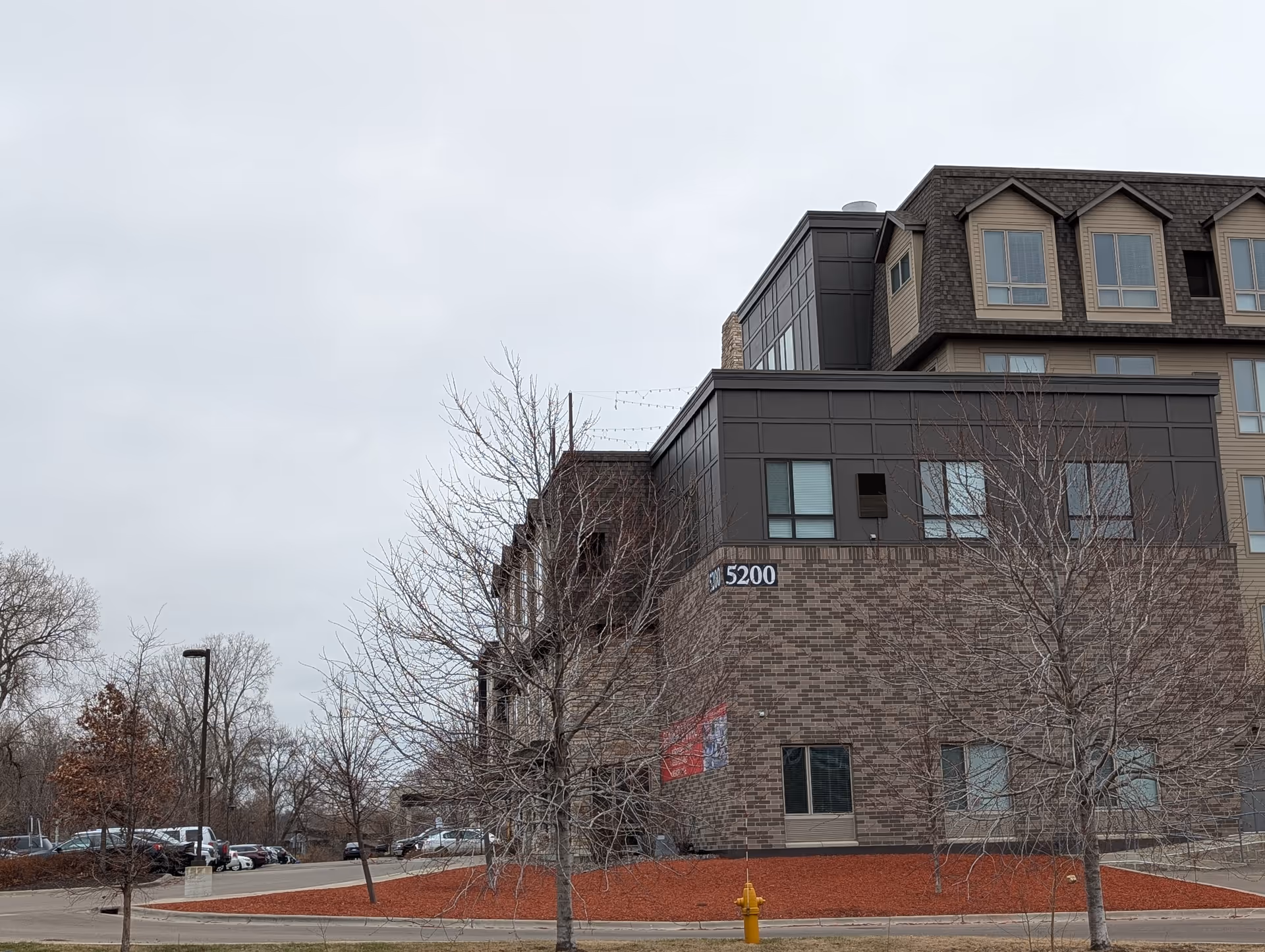 Exterior view of a multi-story senior living facility building with a brick and dark panel facade, leafless trees in front, a parking lot to the left, and a cloudy sky overhead.