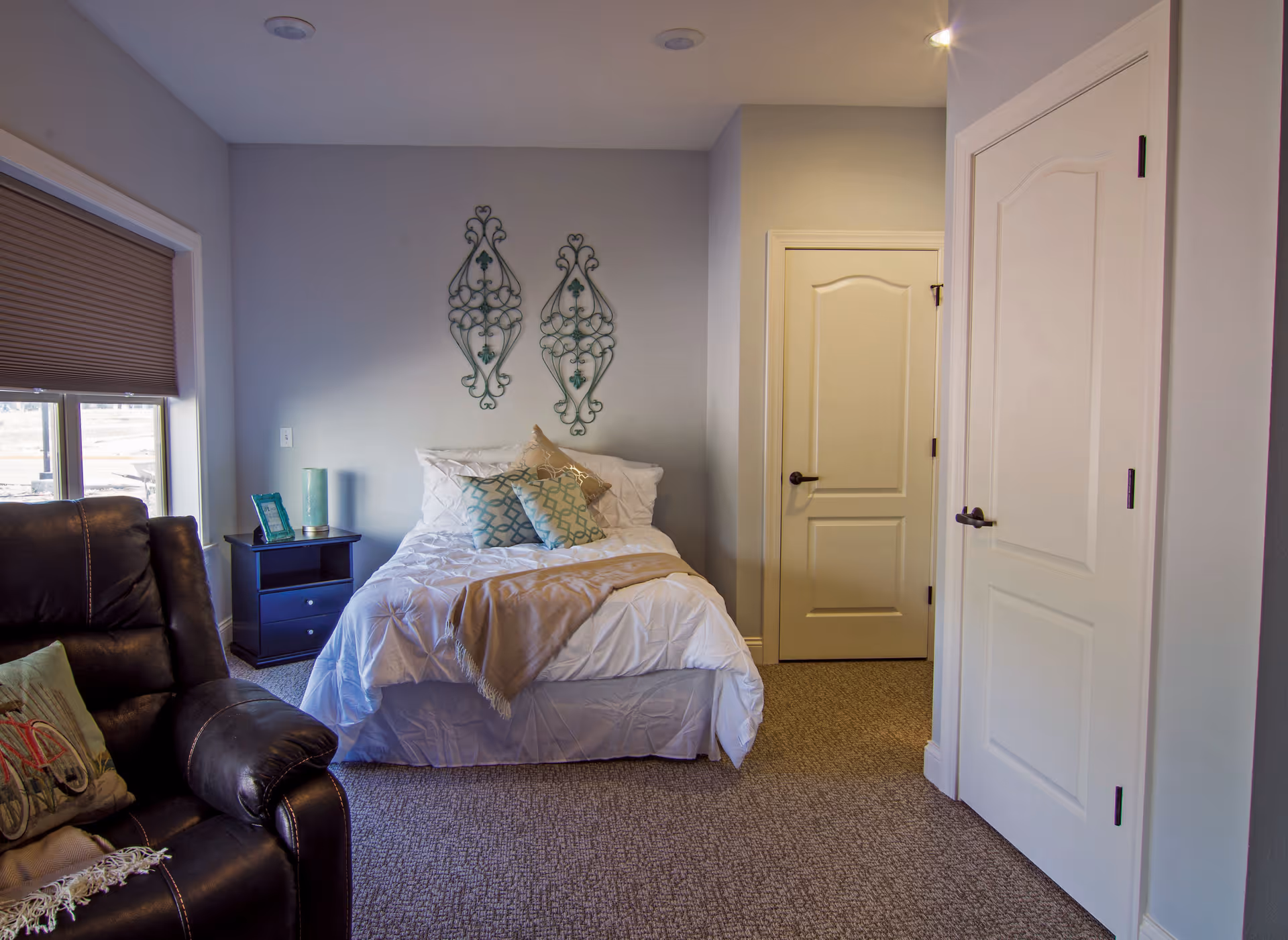 A cozy bedroom with a neatly made bed featuring white bedding and decorative pillows. There is a dark brown leather armchair with a pillow on it to the left, a small black nightstand with a lamp and picture frame next to the bed, and two decorative metal wall hangings above the bed. The room has beige carpet, light gray walls, and two closed white doors on the right side.