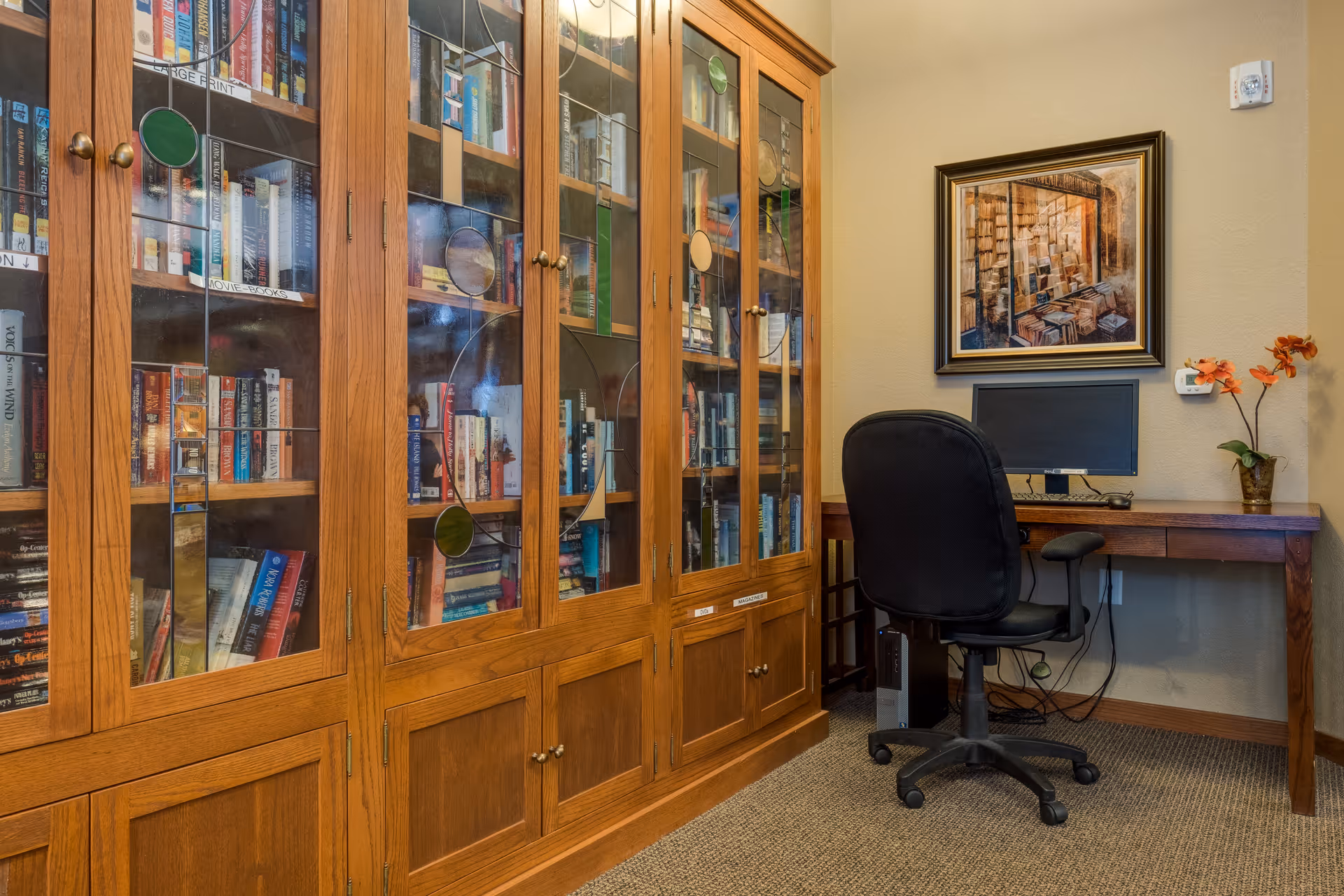 A cozy room featuring a large wooden bookcase with glass doors filled with books, and a wooden desk with a computer monitor and a black office chair. A framed painting hangs on the wall above the desk, and a small vase with orange flowers is placed on the desk.