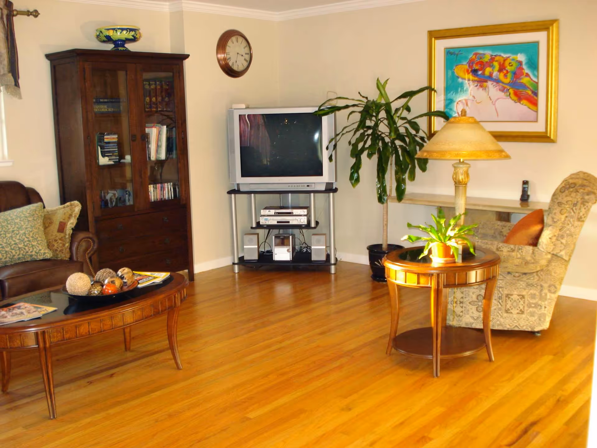 A cozy living room with wooden flooring featuring a brown leather sofa with patterned cushions, a wooden coffee table with decorative items, a wooden cabinet with glass doors holding books and DVDs, a TV on a stand with speakers and electronic devices, a tall green plant, a round wooden side table with a lamp and a small potted plant, an upholstered armchair with a cushion, a wall clock, and a colorful framed painting on the wall.
