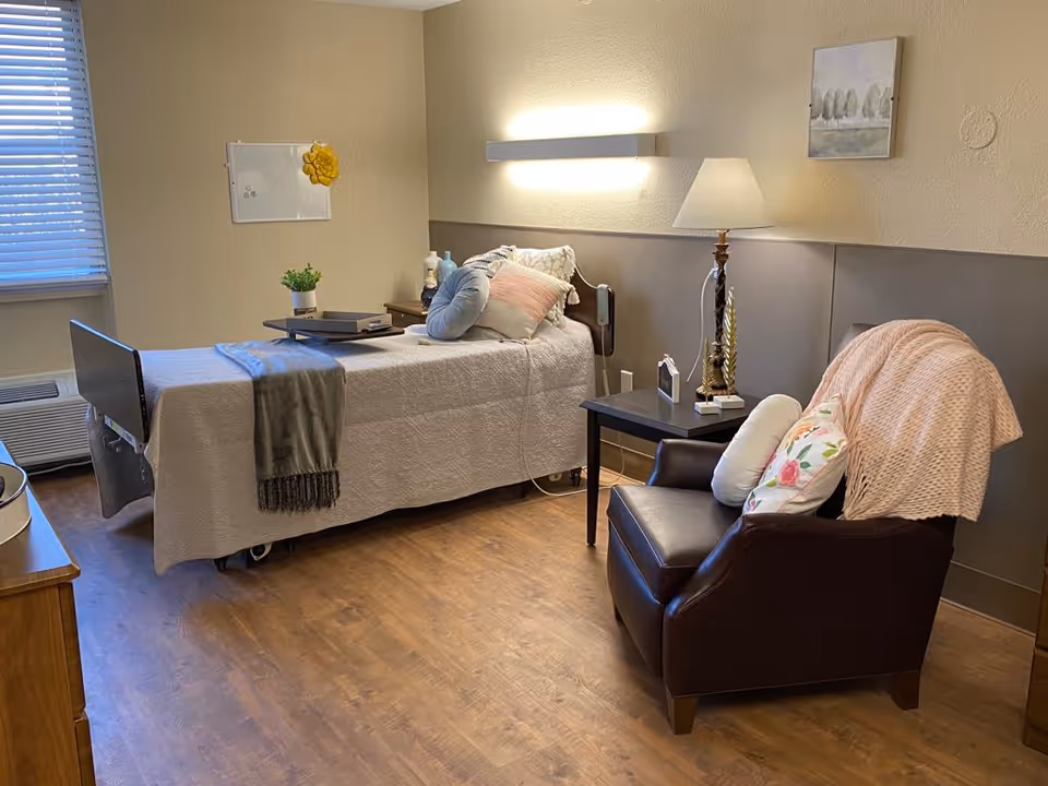 A cozy senior living bedroom with a single bed covered in a beige quilt and multiple pillows. A gray throw blanket is draped over the foot of the bed. Next to the bed is a small wooden nightstand with a lamp, a small clock, and decorative items. A comfortable dark brown armchair with floral and white pillows and a pink throw blanket is positioned nearby. The room has wooden flooring, a window with blinds, and soft lighting from a wall-mounted light fixture and the lamp. A small potted plant and a whiteboard with a yellow flower decoration are on the wall.