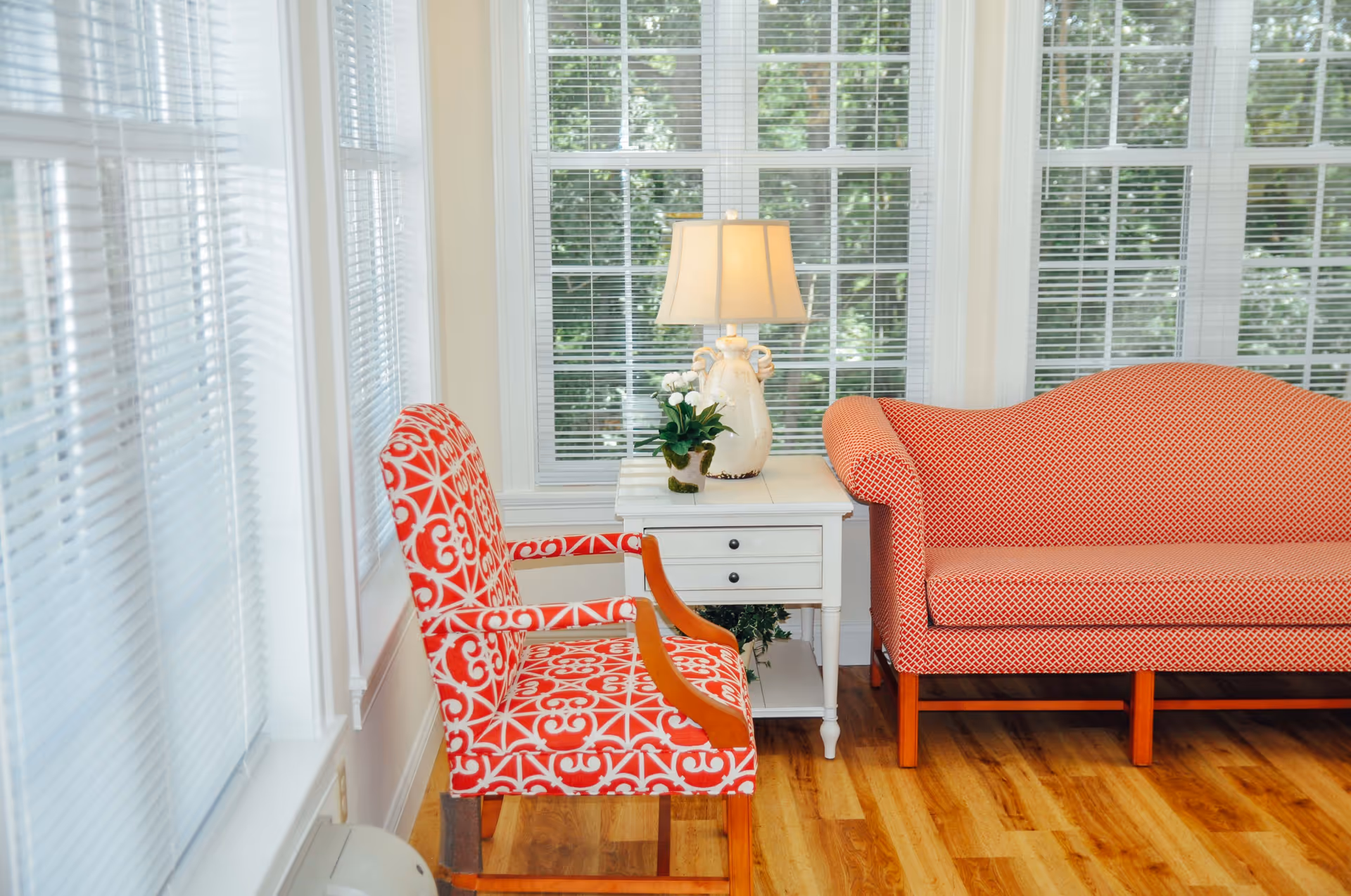 A bright sitting area with a red patterned armchair and a red patterned loveseat. Between them is a white side table with two drawers, holding a decorative lamp and a small potted plant. Large windows with white blinds let in natural light, and the floor is wooden.
