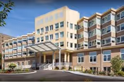 Exterior view of a multi-story senior living facility building with a covered entrance, large windows, and landscaped surroundings under a clear blue sky.