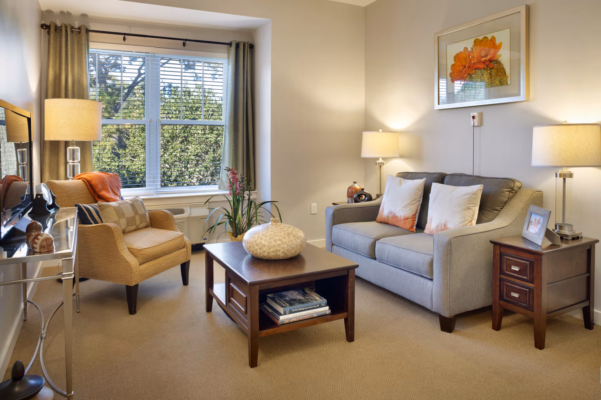 A cozy living room with a gray sofa adorned with two white and orange pillows, a wooden coffee table with a decorative vase, a beige armchair with a striped pillow and orange throw, two table lamps on wooden side tables, a large window with green curtains, and a framed floral artwork on the wall.