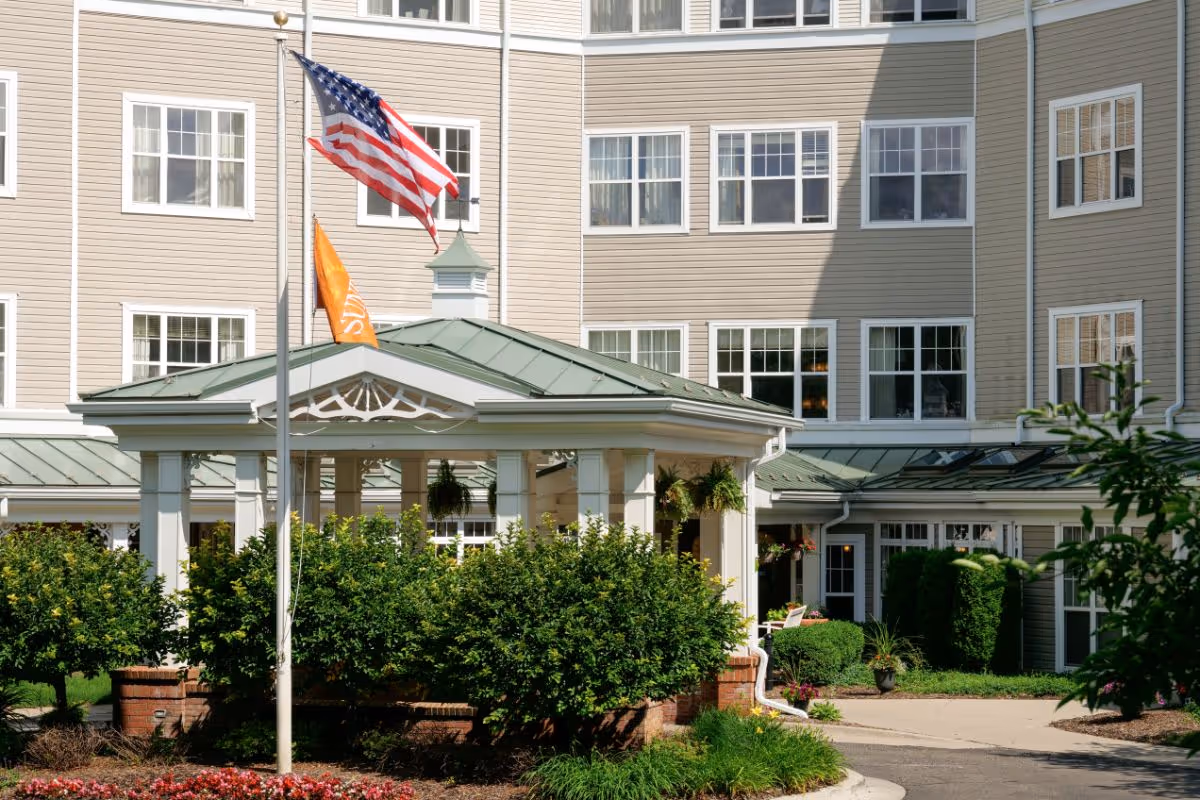 Exterior view of River Oaks Assisted Living & Memory Care building with beige siding and multiple white-framed windows. In front, there is a covered entrance with a green roof, surrounded by green bushes and plants. Two flagpoles display an American flag and an orange flag. The area is well-maintained with a paved driveway and landscaping.