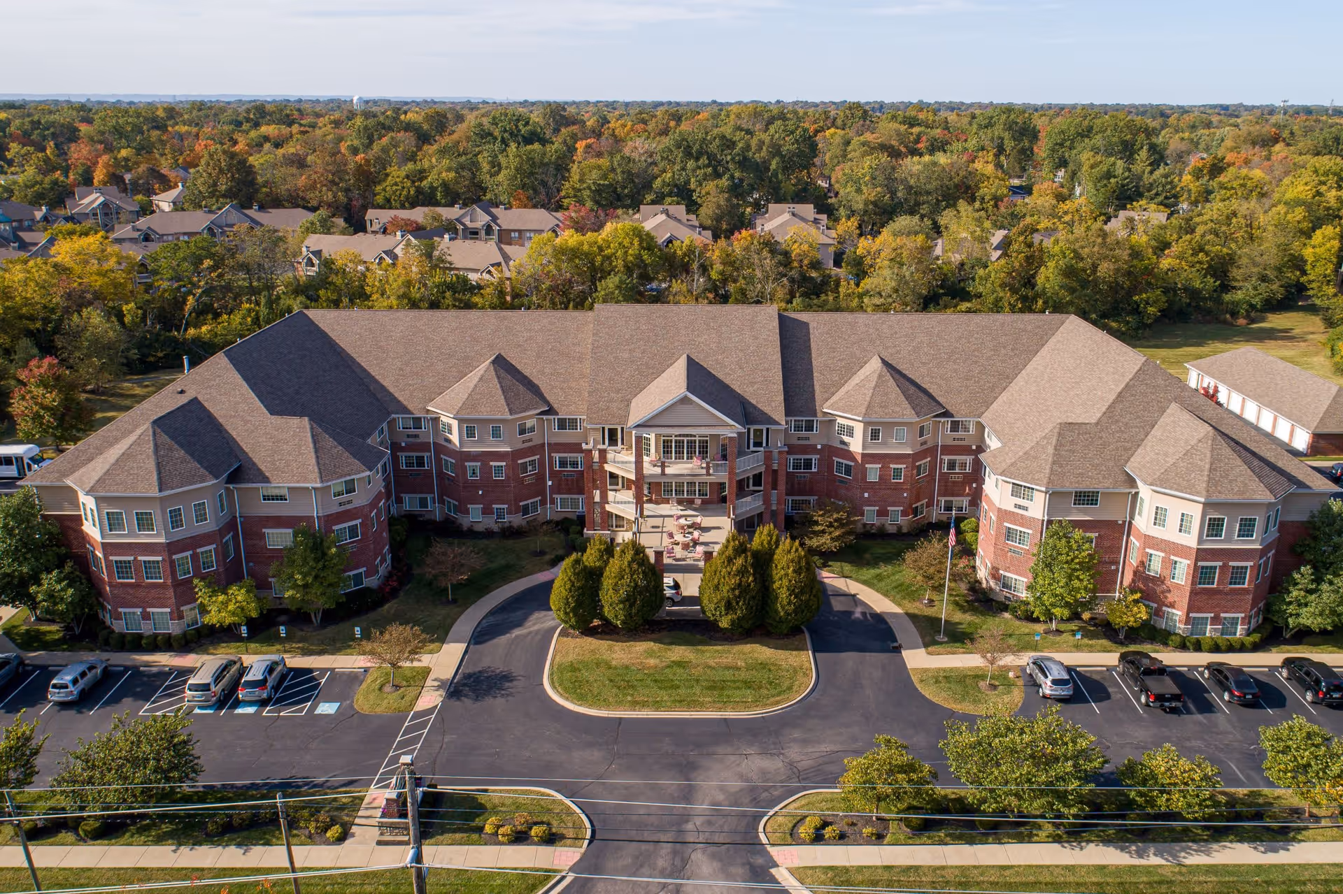 Aerial view of a large senior living facility building with a U-shaped design, surrounded by parking lots and trees with autumn foliage in the background.