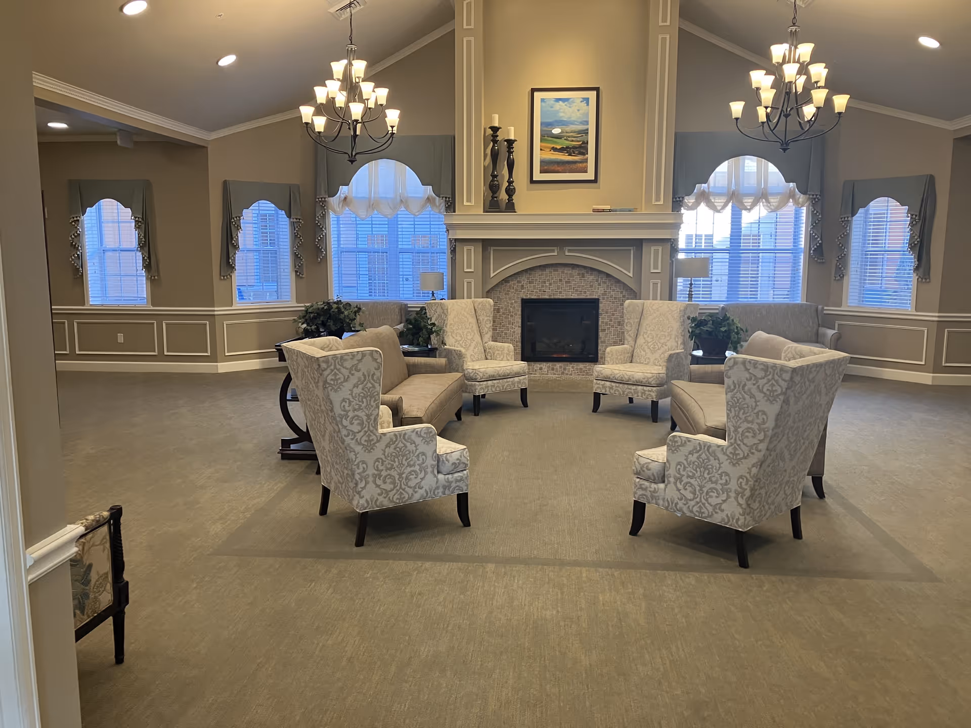 A spacious and elegant living room area with a central fireplace, surrounded by patterned armchairs and sofas arranged in a circle. The room features large windows with decorative curtains, two chandeliers hanging from the ceiling, and a framed landscape painting above the fireplace. The walls have decorative molding and there are plants placed near the windows.