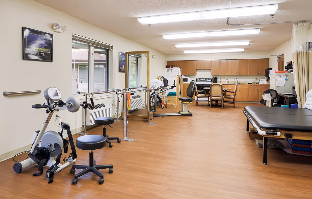A rehabilitation room with exercise equipment including a stationary bike and parallel bars, a large mirror on the wall, a treatment table, and a kitchenette area with cabinets, a stove, microwave, and a table with chairs. The room has wood flooring and fluorescent ceiling lights.