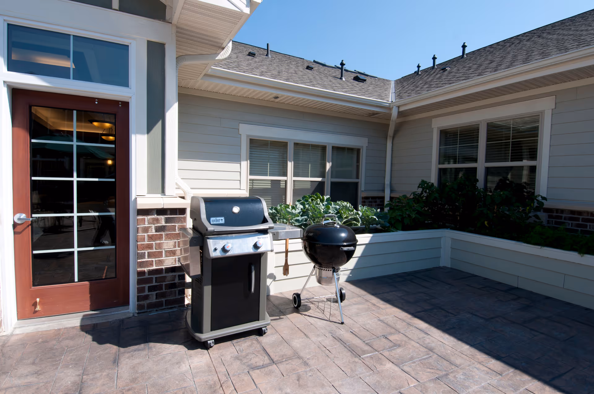 Outdoor patio area with two grills, one rectangular and one round, placed on a tiled floor next to a building with windows and a glass door. Raised garden beds with green plants are visible along the walls under a clear blue sky.