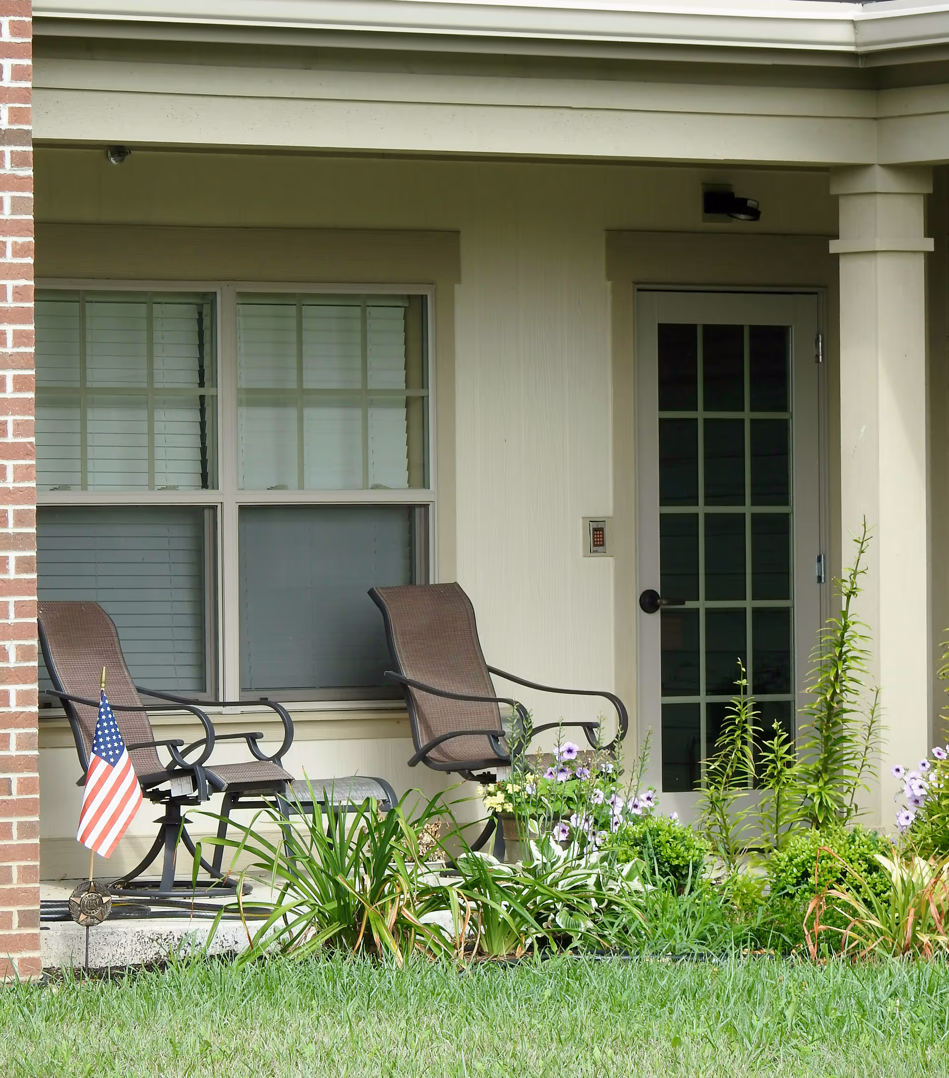 A small outdoor patio area at Logan Acres Senior Community featuring two brown metal chairs with armrests, a small American flag planted in the garden bed, various green plants and purple flowers, a window with closed blinds, and a door with multiple glass panes.