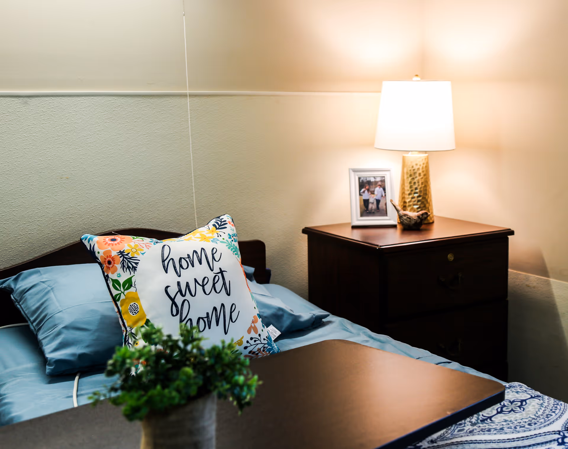 A cozy bedroom scene with a bed featuring blue sheets and a decorative pillow that says 'home sweet home'. Next to the bed is a wooden nightstand with a lit lamp, a framed photo, and a small decorative bird. In the foreground, there is a small green potted plant.