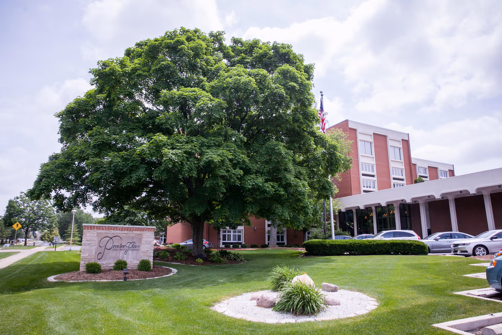 Exterior view of Proctor Place Senior Living facility showing a large green tree, a manicured lawn with landscaping, a stone sign with the facility name, and a multi-story brick building with parked cars in front under a partly cloudy sky.