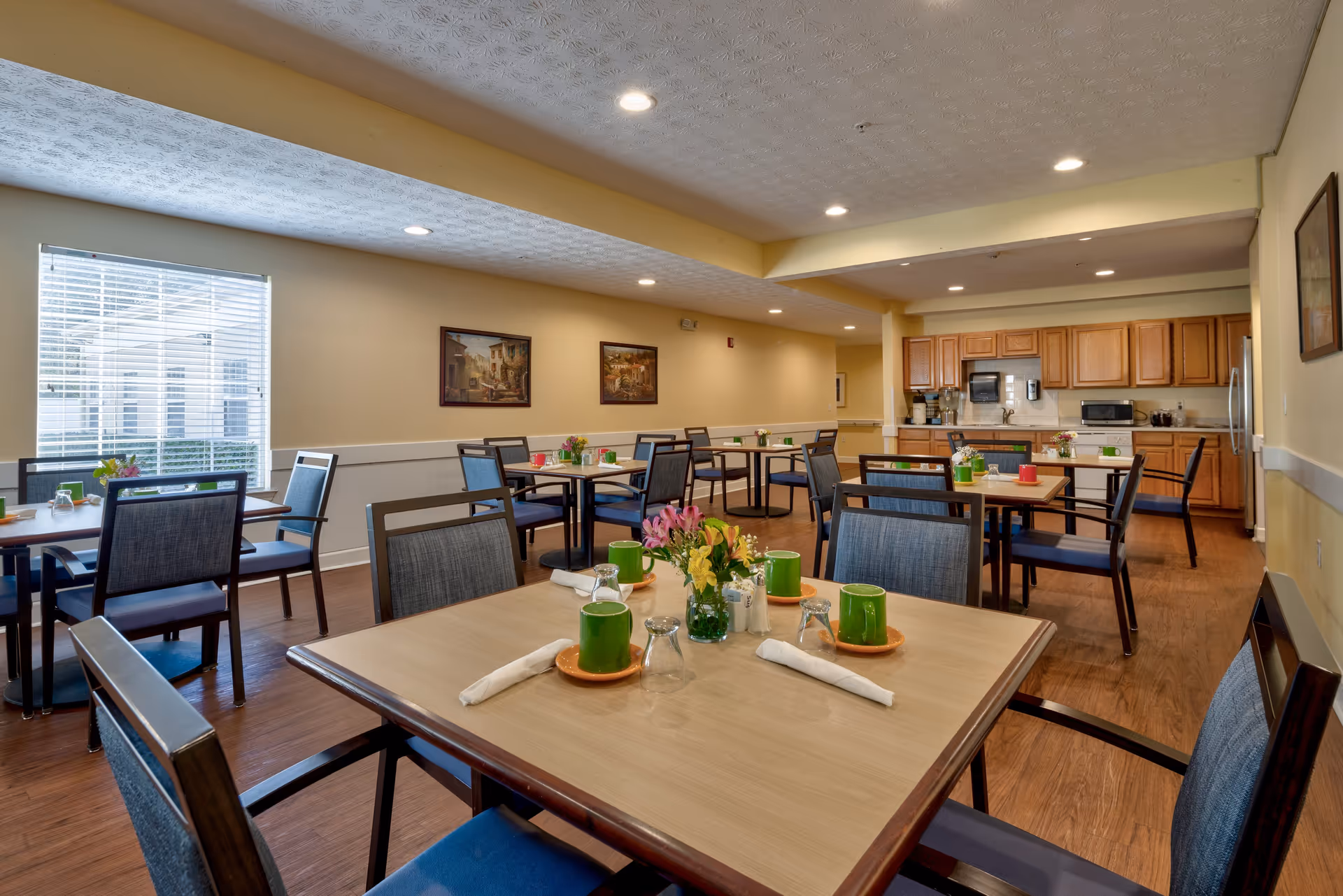 A dining room in a senior living facility with several tables and chairs arranged neatly. Each table is set with green mugs, glasses, napkins, and small flower arrangements. The room has wooden flooring, beige walls, recessed ceiling lights, and a window with blinds letting in natural light. In the background, there is a kitchenette area with wooden cabinets, a microwave, and a refrigerator.