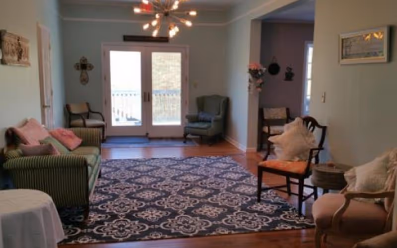 A cozy living room with a patterned area rug covering the wooden floor. The room features a green striped sofa with pink cushions on the left, a green armchair near double glass doors leading outside, and several wooden chairs with cushions on the right. The walls are painted light blue, and there is a modern chandelier hanging from the ceiling. Decorative items such as wall art and flowers are also visible.