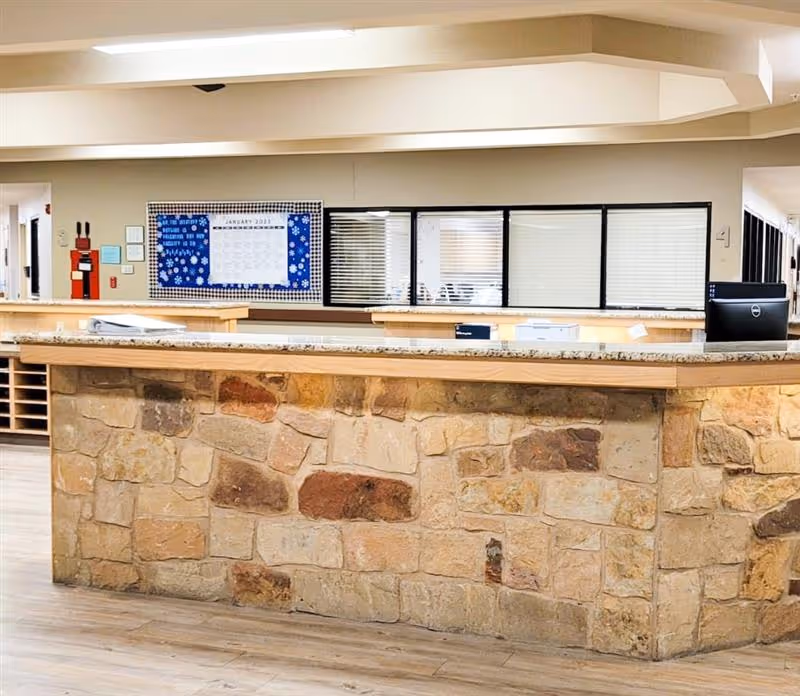 Reception desk area inside a senior living facility with a stone front counter and a granite countertop. Behind the desk are windows with blinds and a bulletin board with a calendar and notices on the wall.
