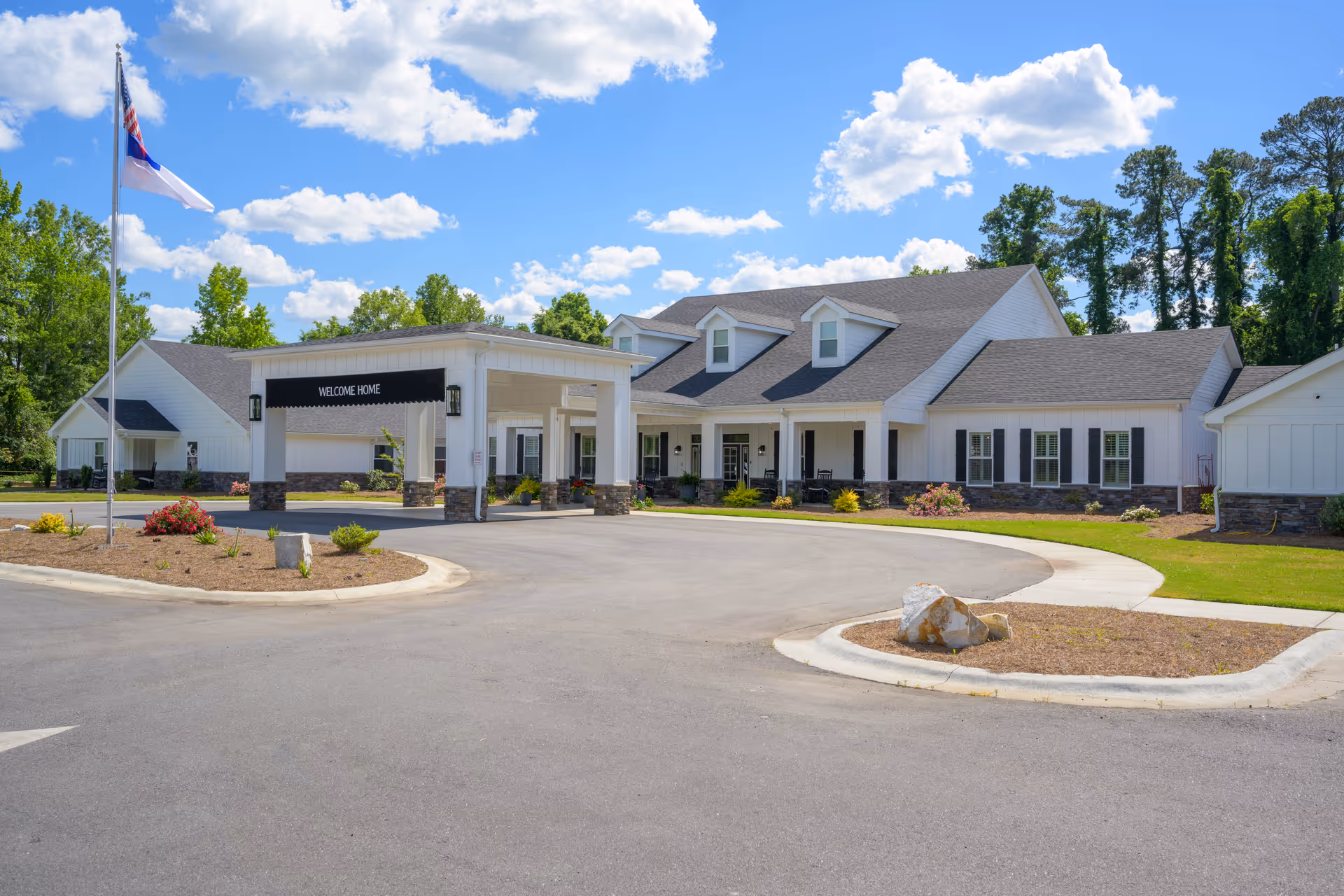 Front entrance of a single-story white senior living building with a covered porte-cochère reading "WELCOME HOME" and a flagpole in front.