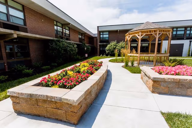 Outdoor courtyard area at The Village at Olathe featuring raised stone flower beds with colorful flowers, a concrete walkway, and a wooden gazebo. The courtyard is surrounded by a two-story brick building with multiple windows.