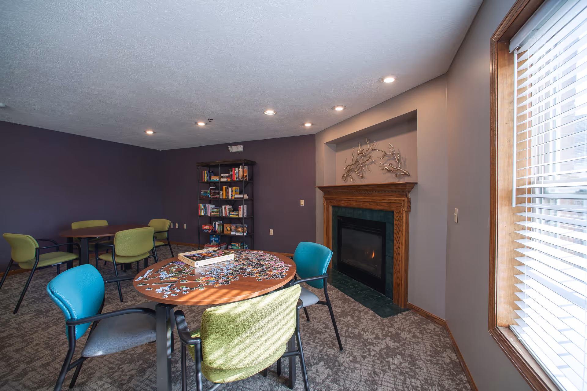 Small communal lounge with round tables and chairs, a bookshelf, and a fireplace under recessed lighting.