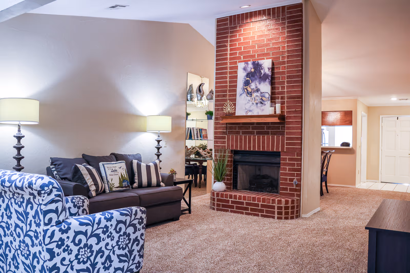 Cozy living room with a patterned armchair, dark sofa with pillows, twin lamps, and a red brick fireplace.