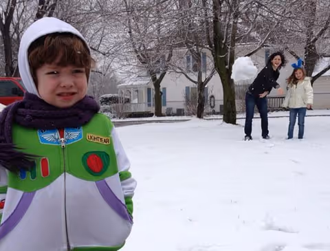 A child dressed in a Buzz Lightyear costume stands in the snow in the foreground, while a woman and another child in winter clothing are in the background playing with a snowball in a snowy residential area with houses and trees.