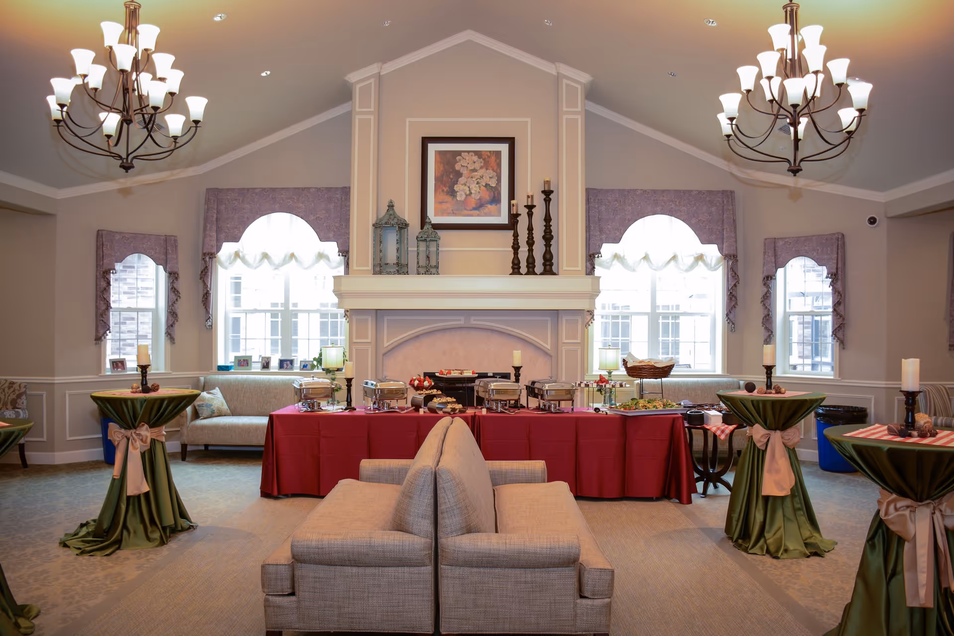 A spacious, elegant common area with two chandeliers hanging from a vaulted ceiling. The room features a central fireplace with decorative candles and a framed floral painting above it. In front of the fireplace, a long table covered with a red tablecloth holds several chafing dishes and food trays. Two beige sofas face each other in the foreground. The room has large windows with purple valances, and there are three tall cocktail tables draped with green cloth and tied with beige ribbons, each holding a candle and small decorative items.