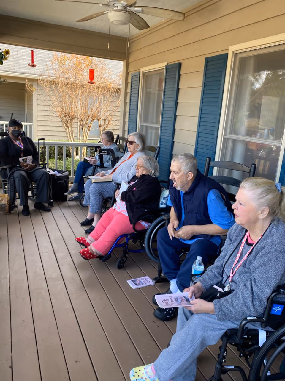 A group of elderly people sitting on a covered wooden porch outside a building. Some are in wheelchairs and others are seated on chairs. They appear to be engaged in an activity, holding papers or booklets. The porch has a ceiling fan and blue shutters on the windows of the building.